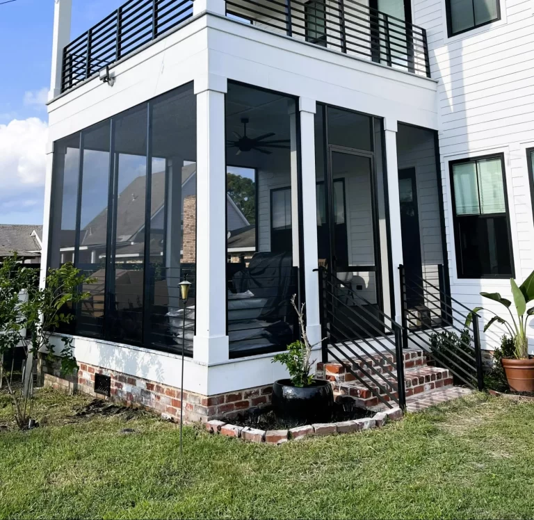 Modern white house with a screened porch featuring black railings and stairs. There's a small yard with a curved brick edging and potted plants. A ceiling fan is visible inside the porch.