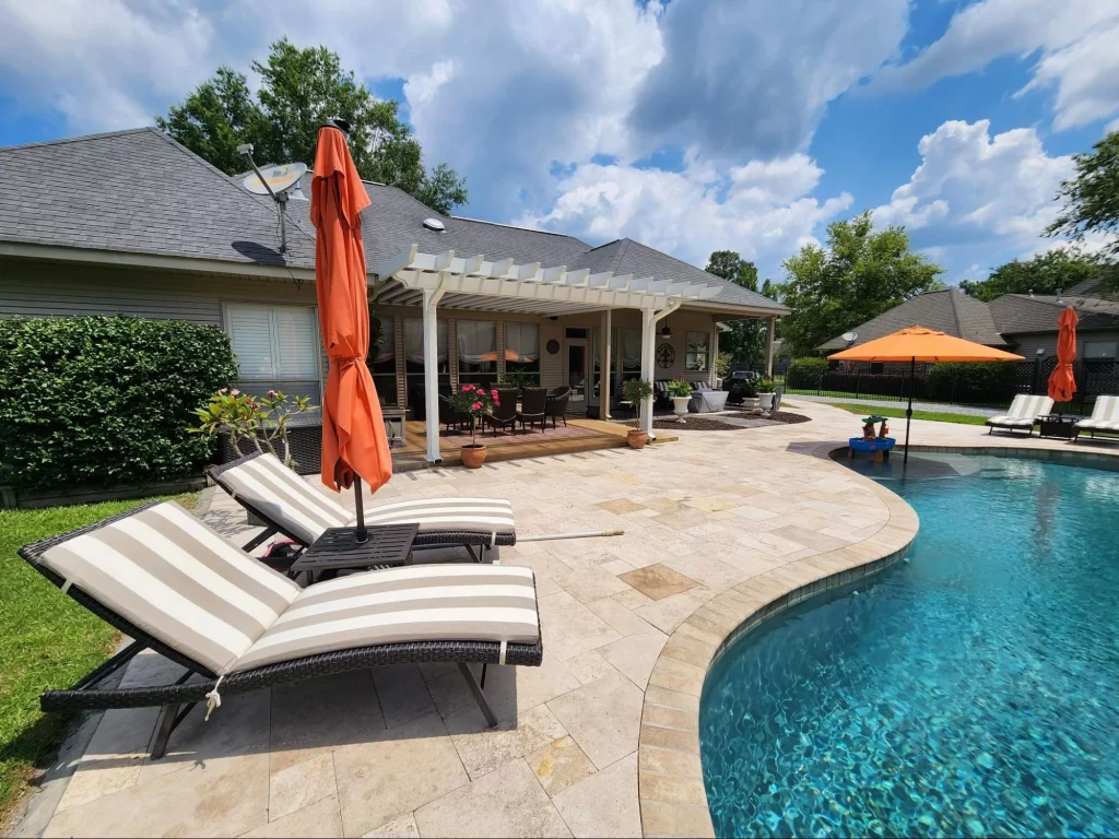 Backyard view of a house with a patio, pergola, and pool. Two striped lounge chairs and orange umbrellas are placed beside the pool. Sky is partly cloudy.