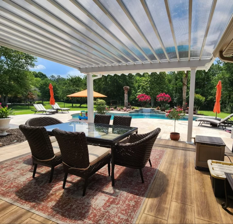 A covered patio with a glass table and wicker chairs overlooks a pool area with lounge chairs, orange umbrellas, and landscaped greenery.